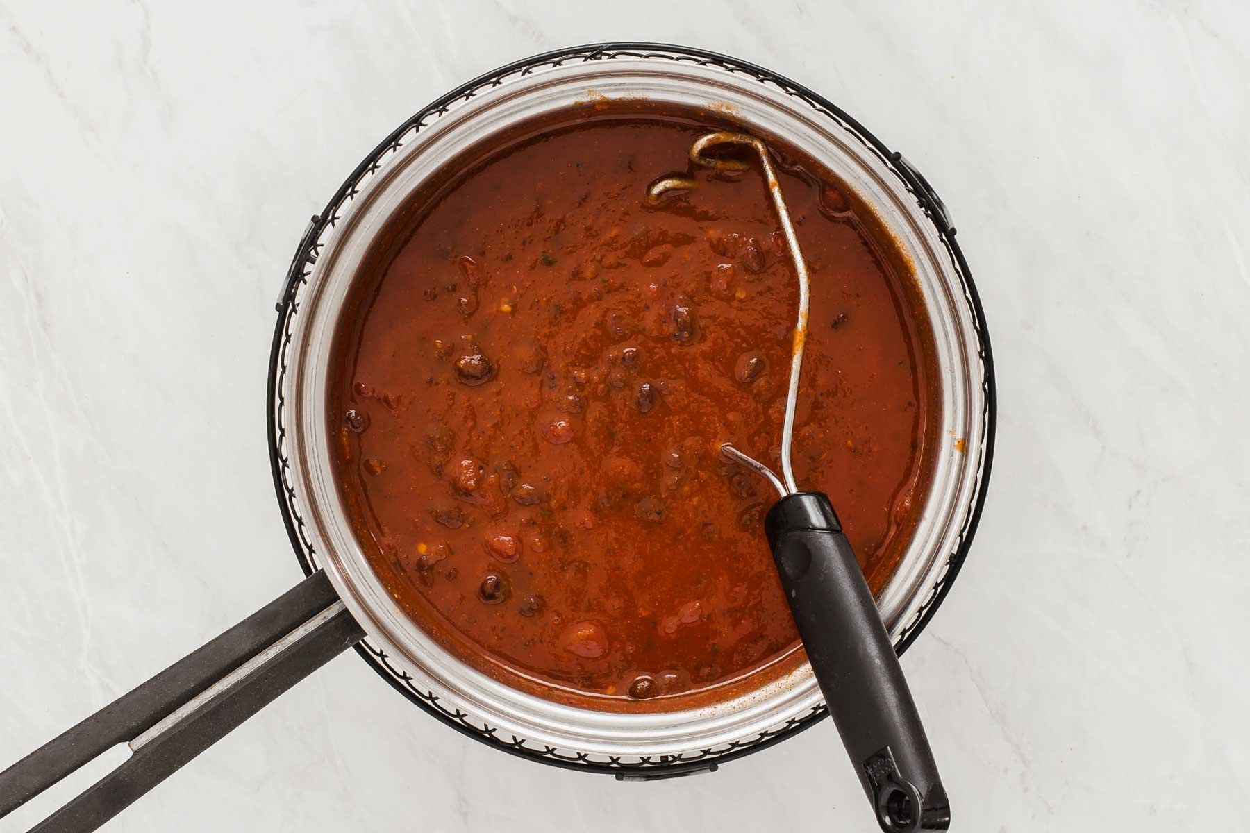 Red tomato sauce in a silver pot with black beans and a potato masher.