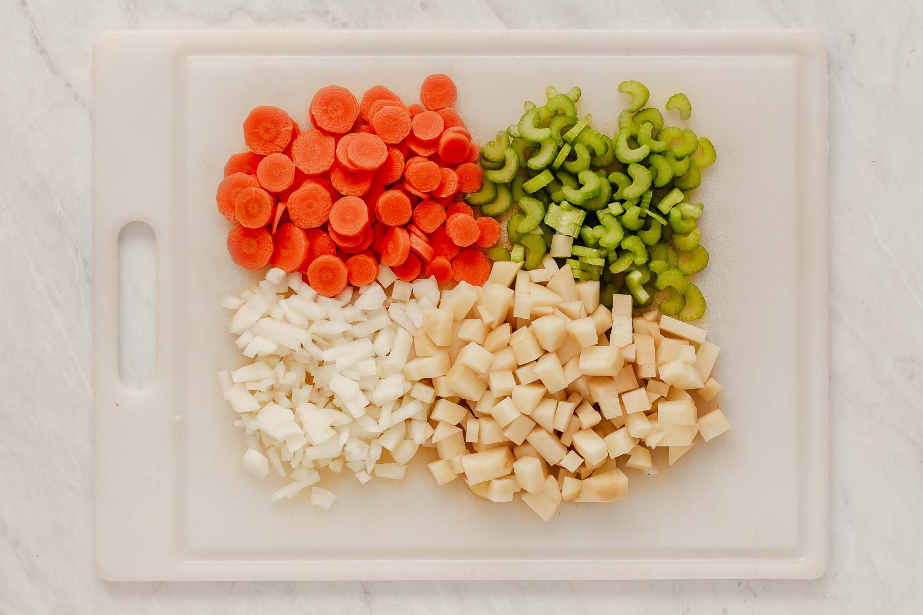 Piles of chopped carrots, celery, onions and potatoes on white cutting board.