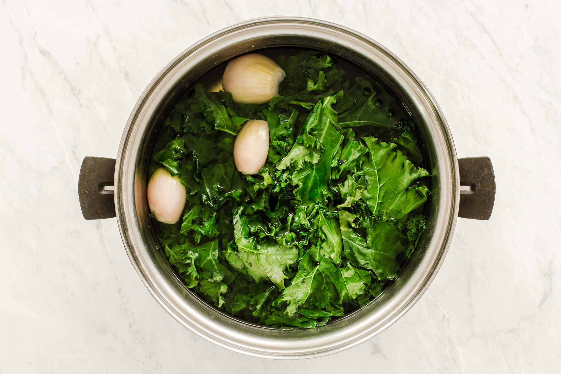 Silver pot with water that has garlic, shallots and kale.