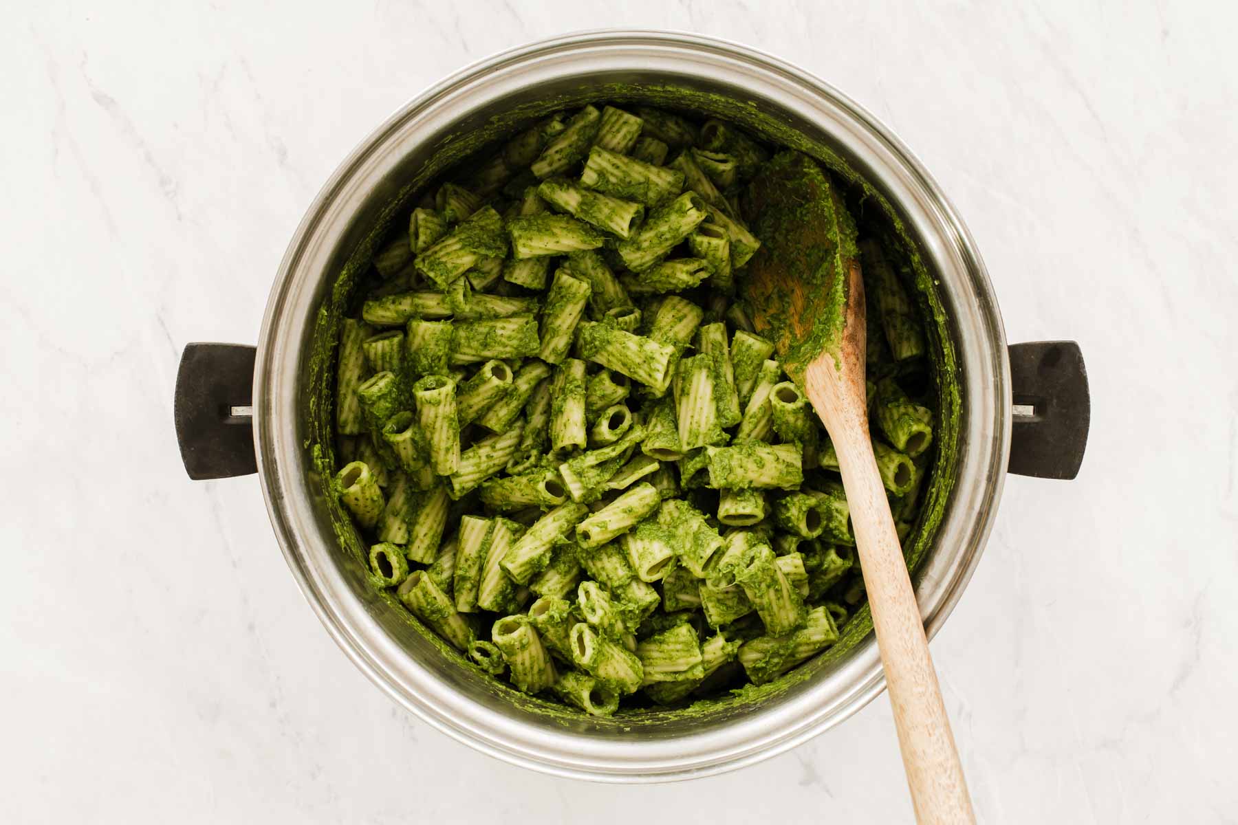 Bright green kale pesto mixed with rigatoni pasta in silver pot.