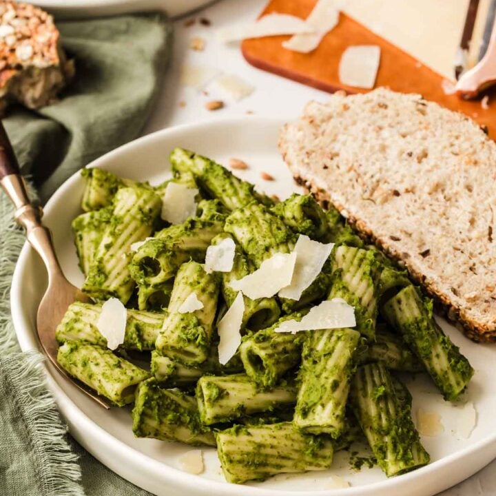 Vertical image of kale pesto on rigatoni pasta with sliced bread on side.
