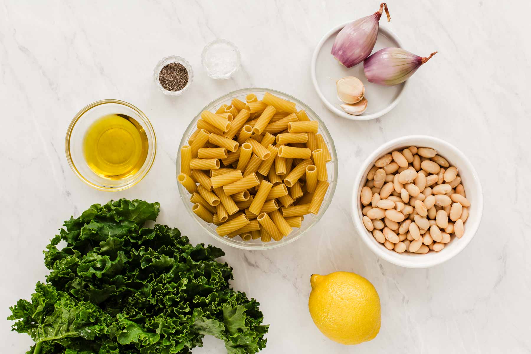 Greens, pasta, beans, and shallots in small bowls on marble counter.