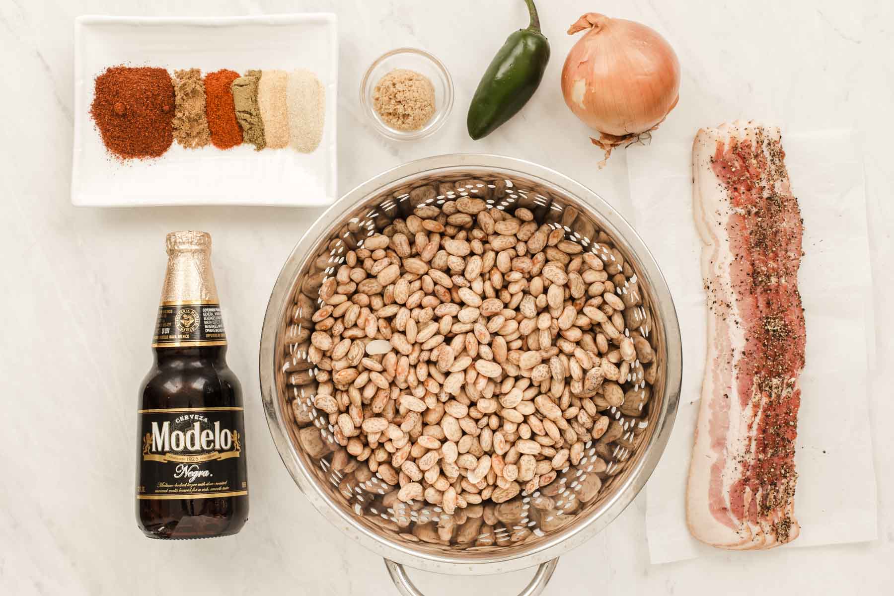 Bowl of soaked pinto beans, bottle of beer, spices and bacon on white table.