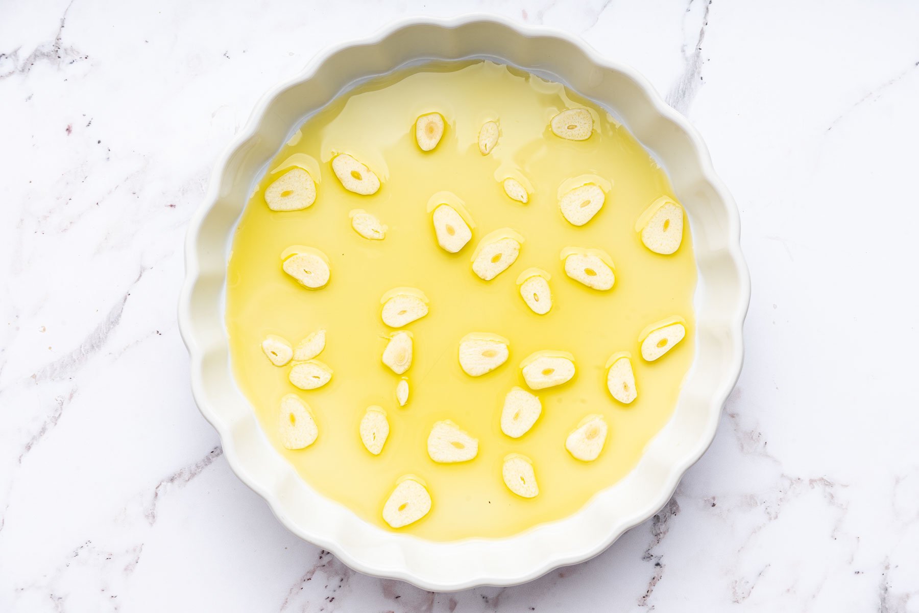 Round white pie dish with a layer of olive oil and thinly sliced garlic cloves.