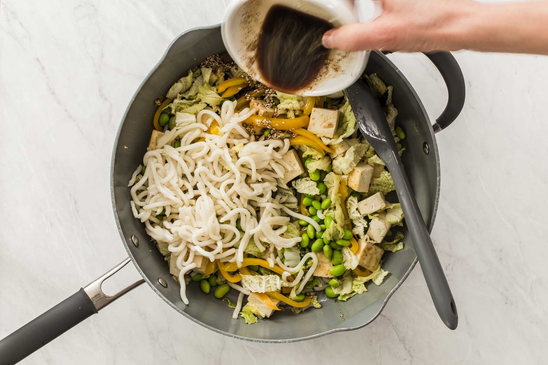 Hand pouring stir fry sauce over noodles and vegetables in grey pan.