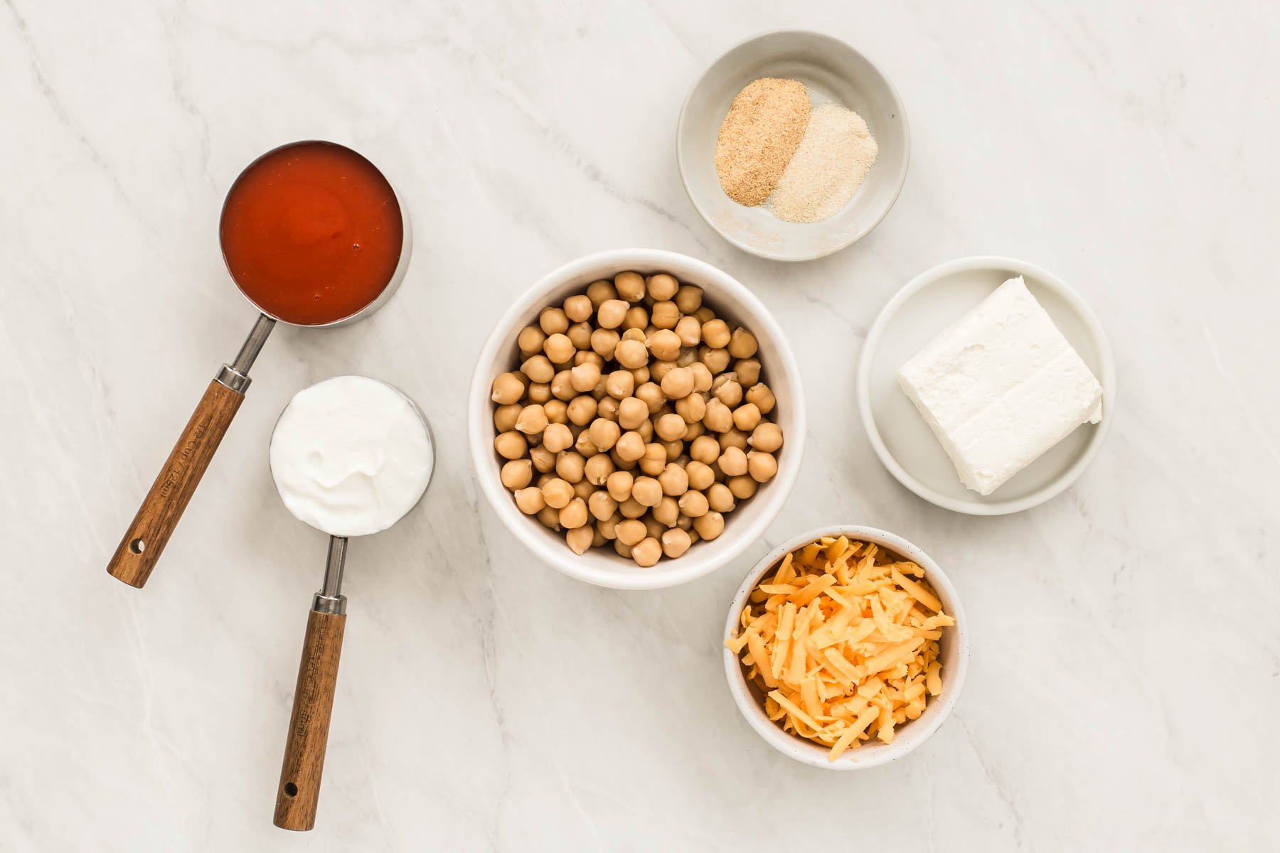 Measuring cups with hot sauce, sour cream, cream cheese and a bowl of chickpeas.